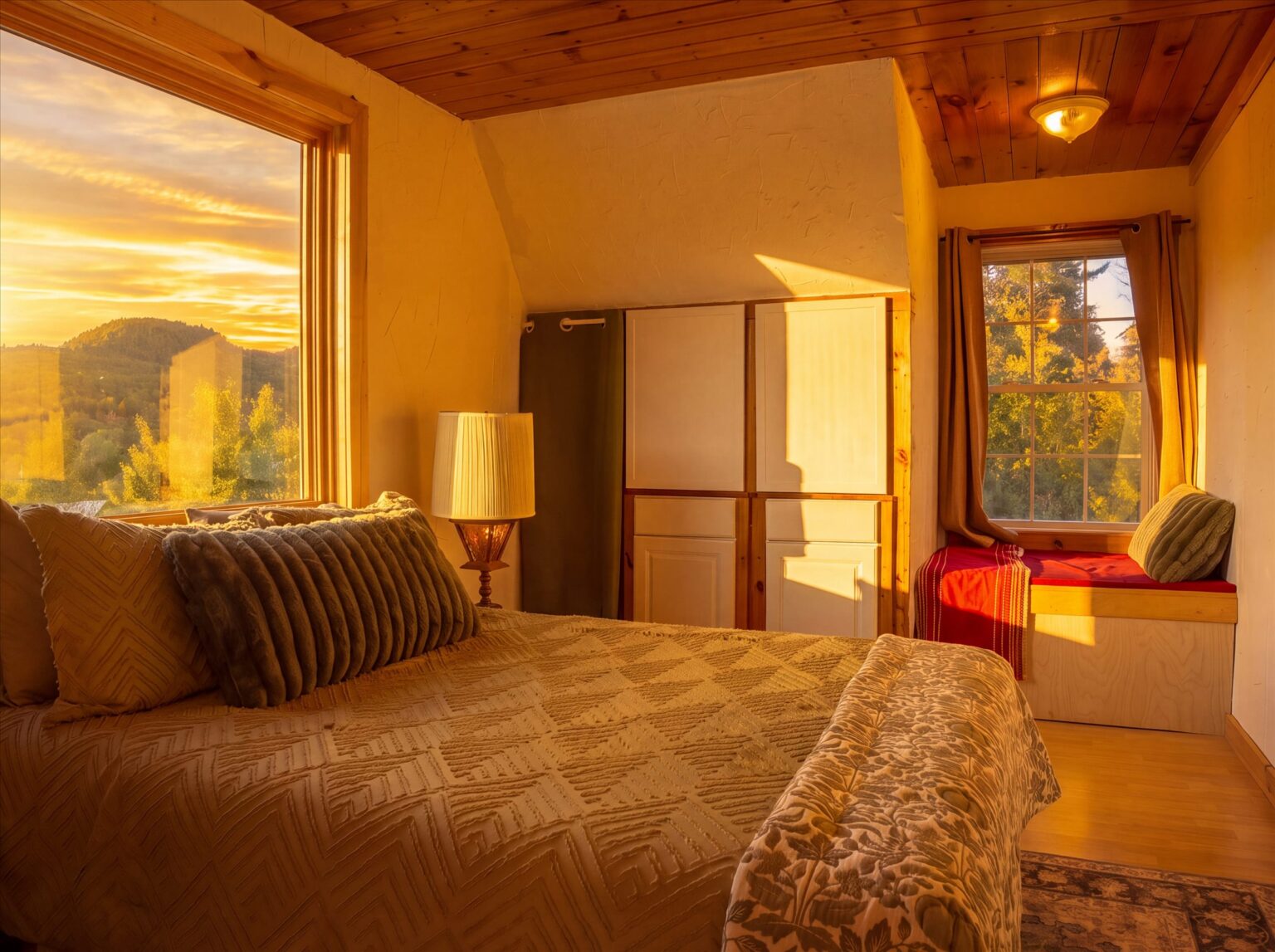 Bedroom in golden light with a bed and tan bedspread beside a large window, a wardrobe, and a small window seat with a red cushion