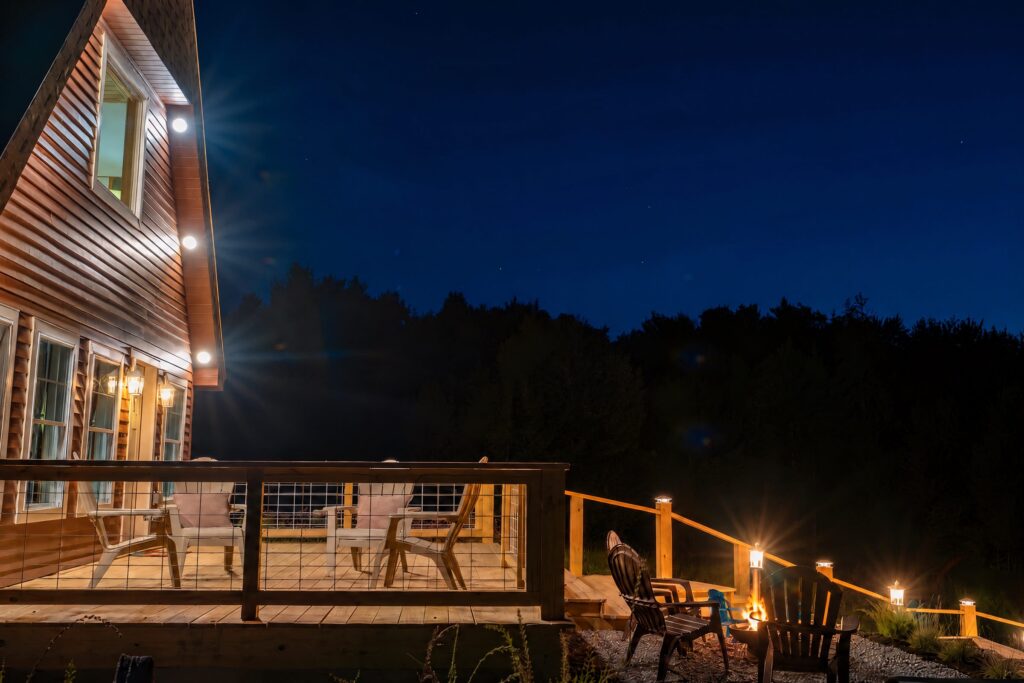 Cabin deck at night with Adirondack chairs, a lit cabin wall and a glowing fire pit in the foreground