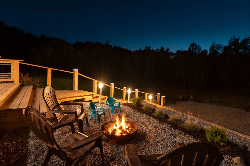 Fire pit with flames, Adirondack chairs and lit railing posts beside the deck at night