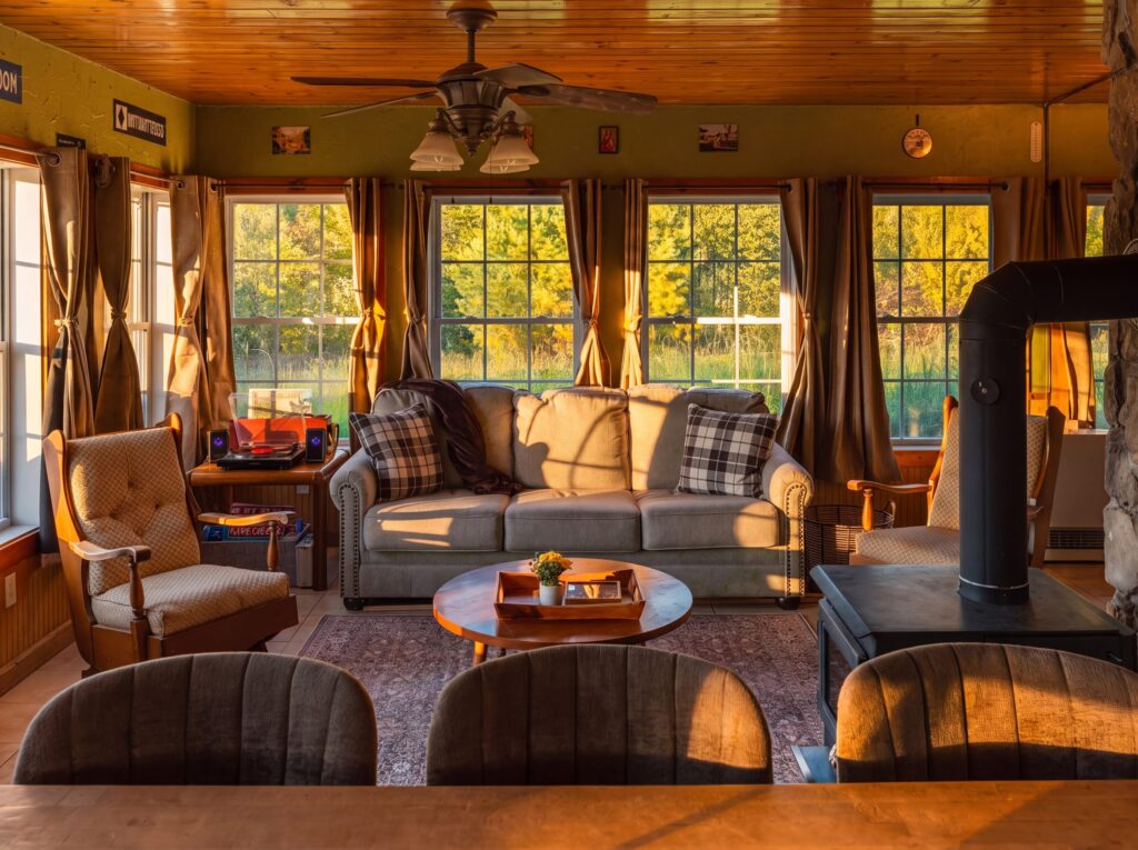 Living room seen across a wooden table, with a gray sofa beneath a row of windows, wooden armchairs, a black wood stove and a stone wall
