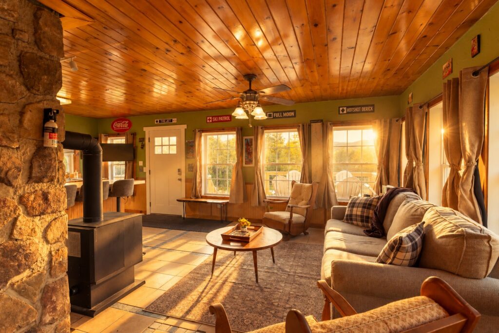 Living room with a stone wall and black wood stove at left, a round wooden coffee table, and a gray sofa with plaid pillows beneath tall windows, under a pine-paneled ceiling in golden light