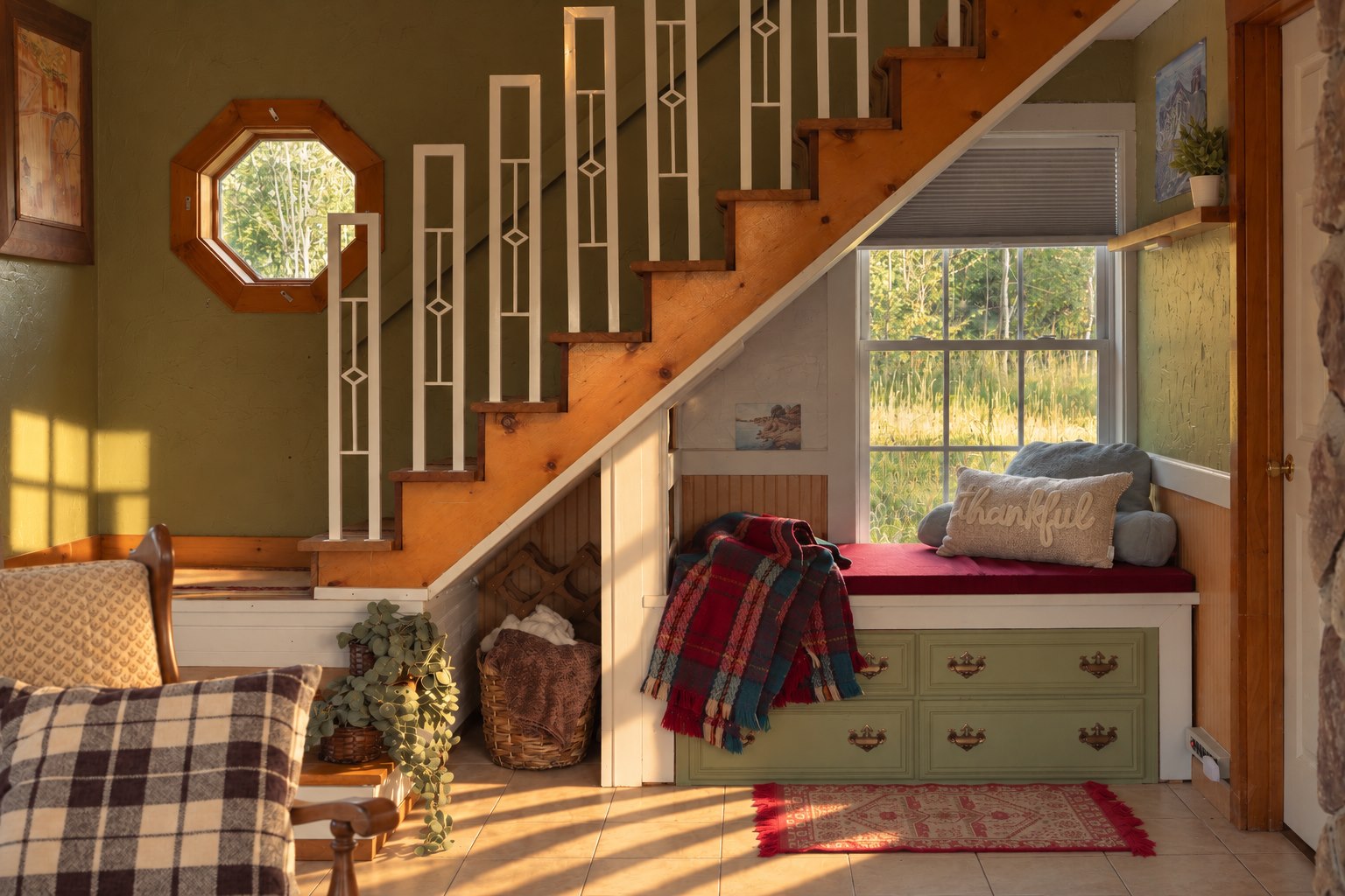 Reading nook beneath a wooden staircase: a window seat with a red cushion and plaid throw above a green drawer chest, beside a window overlooking tall grass