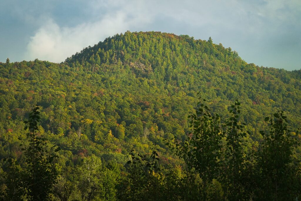 Forested hill with a rocky patch near the summit under a cloudy sky, with early autumn color