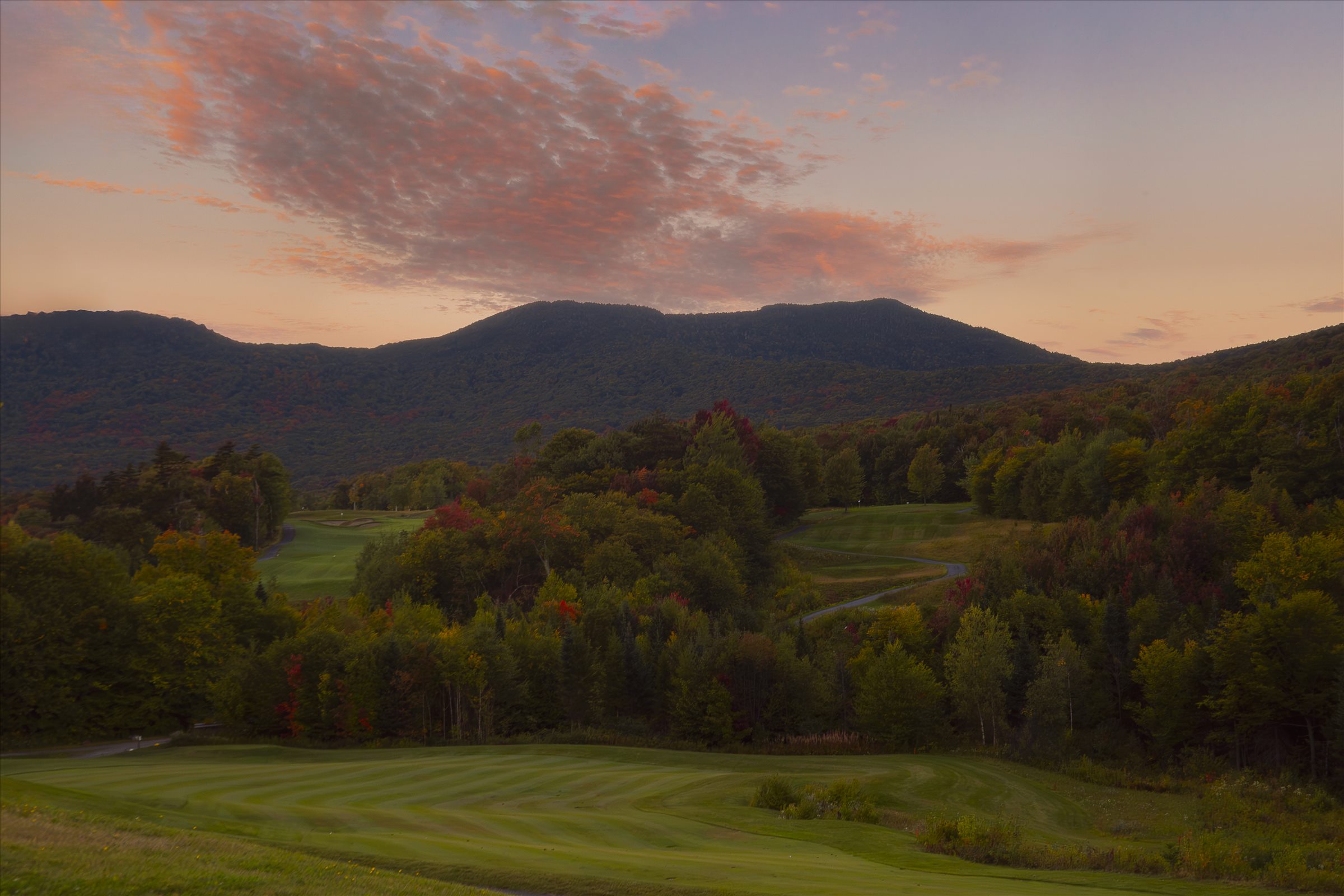 Jay Peak golf course fairways beside autumn-colored woods, with mountains and a pink-tinged sunset sky beyond