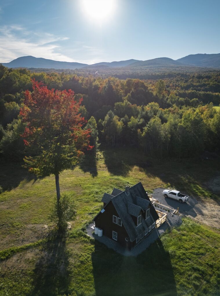 Aerial view of the cabin and a red maple in a clearing, with forest and mountains beyond