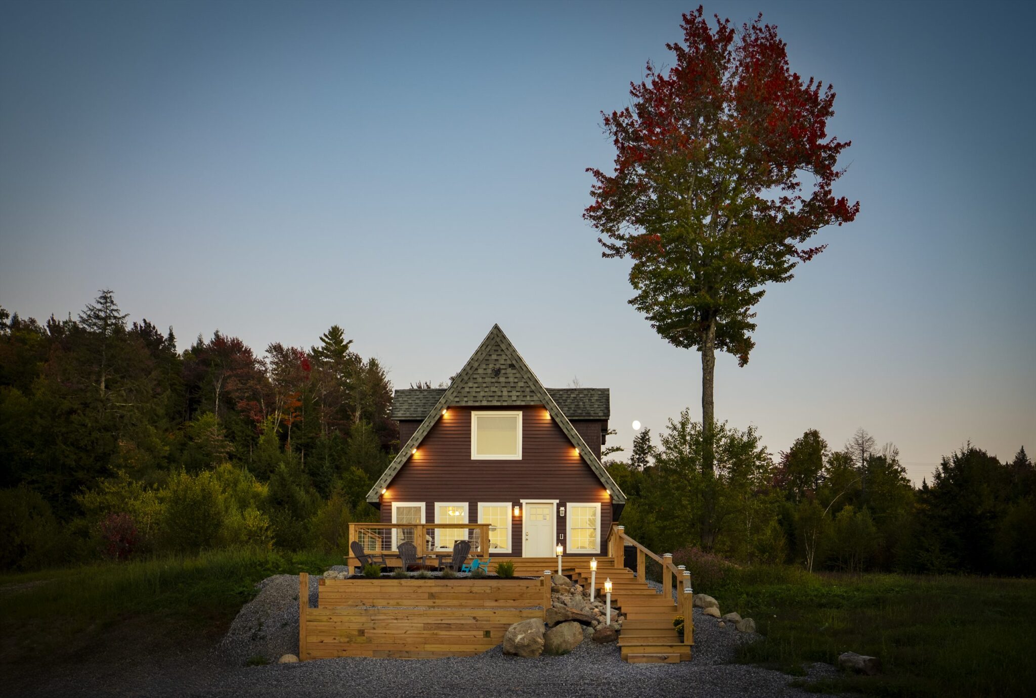 Steep-gabled brown cabin with lit windows and a wooden deck at dusk, beside a tall maple with red leaves