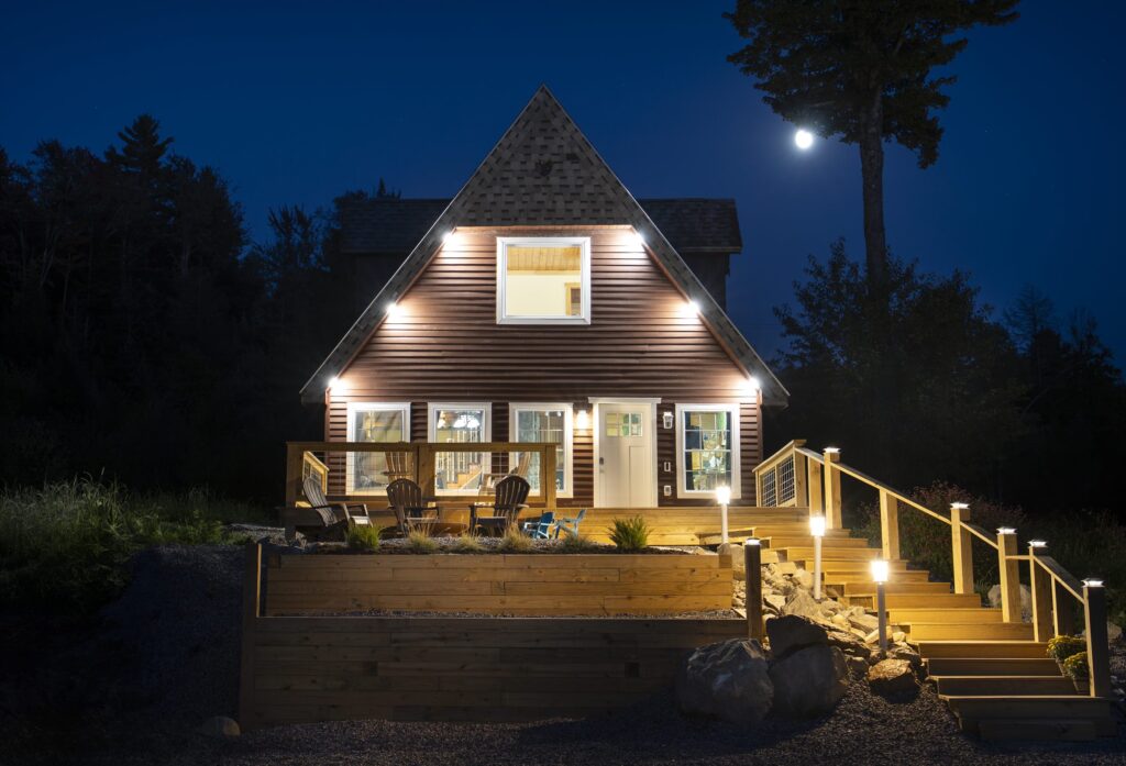 Cabin with warmly lit windows and deck stairs under a night sky with a bright moon