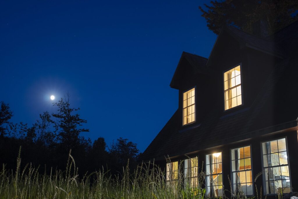 Cabin windows glowing against a deep blue night sky with the moon above the trees