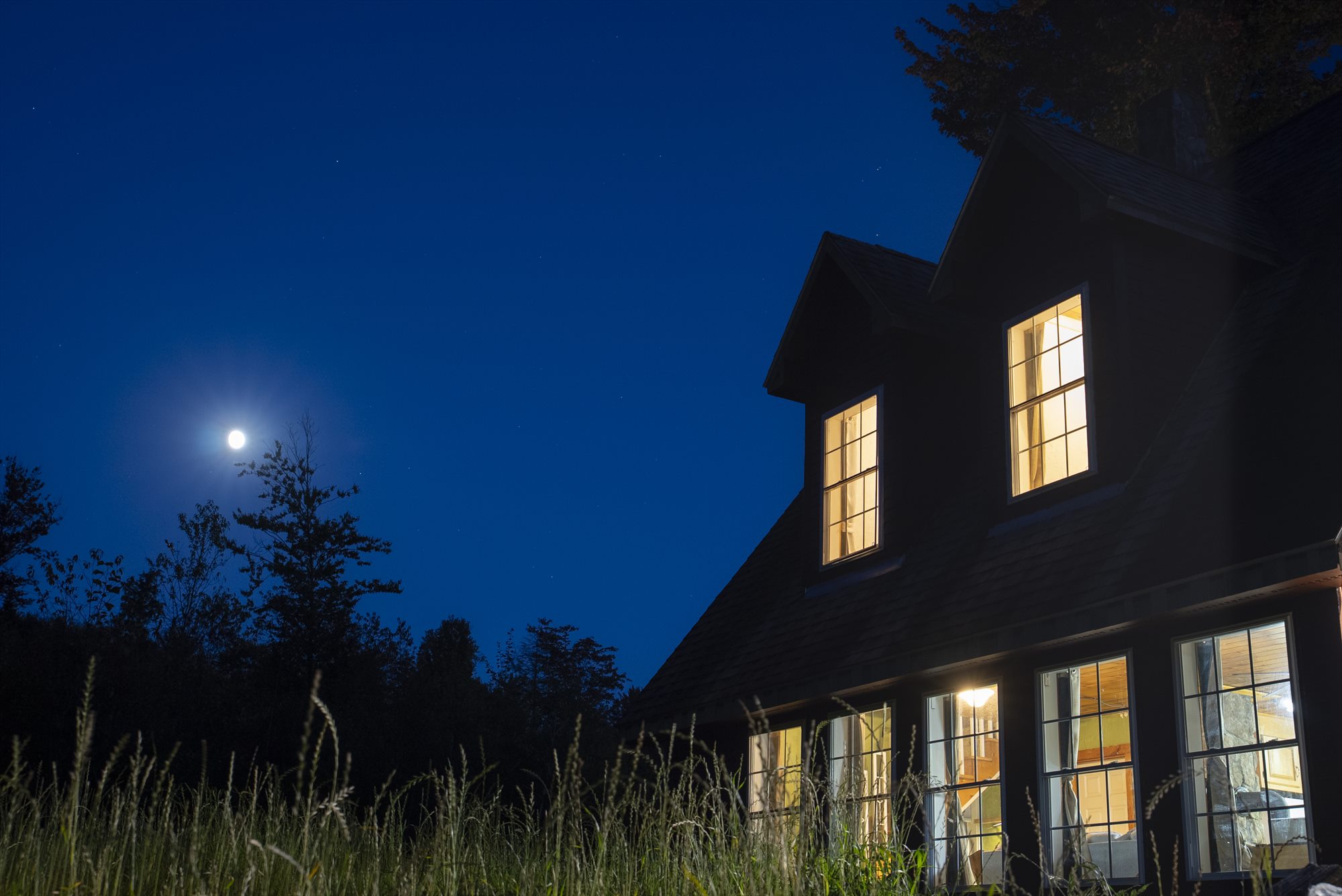 Cabin windows glowing against a deep blue night sky with the moon above the trees