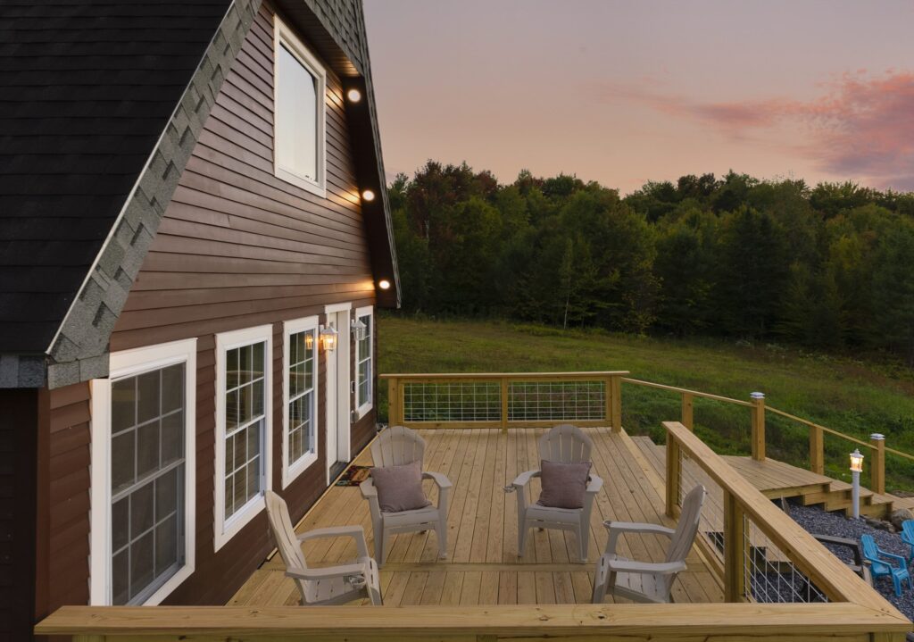 Wooden deck with four Adirondack chairs and a wire-panel railing at dusk, wall lights glowing on the siding