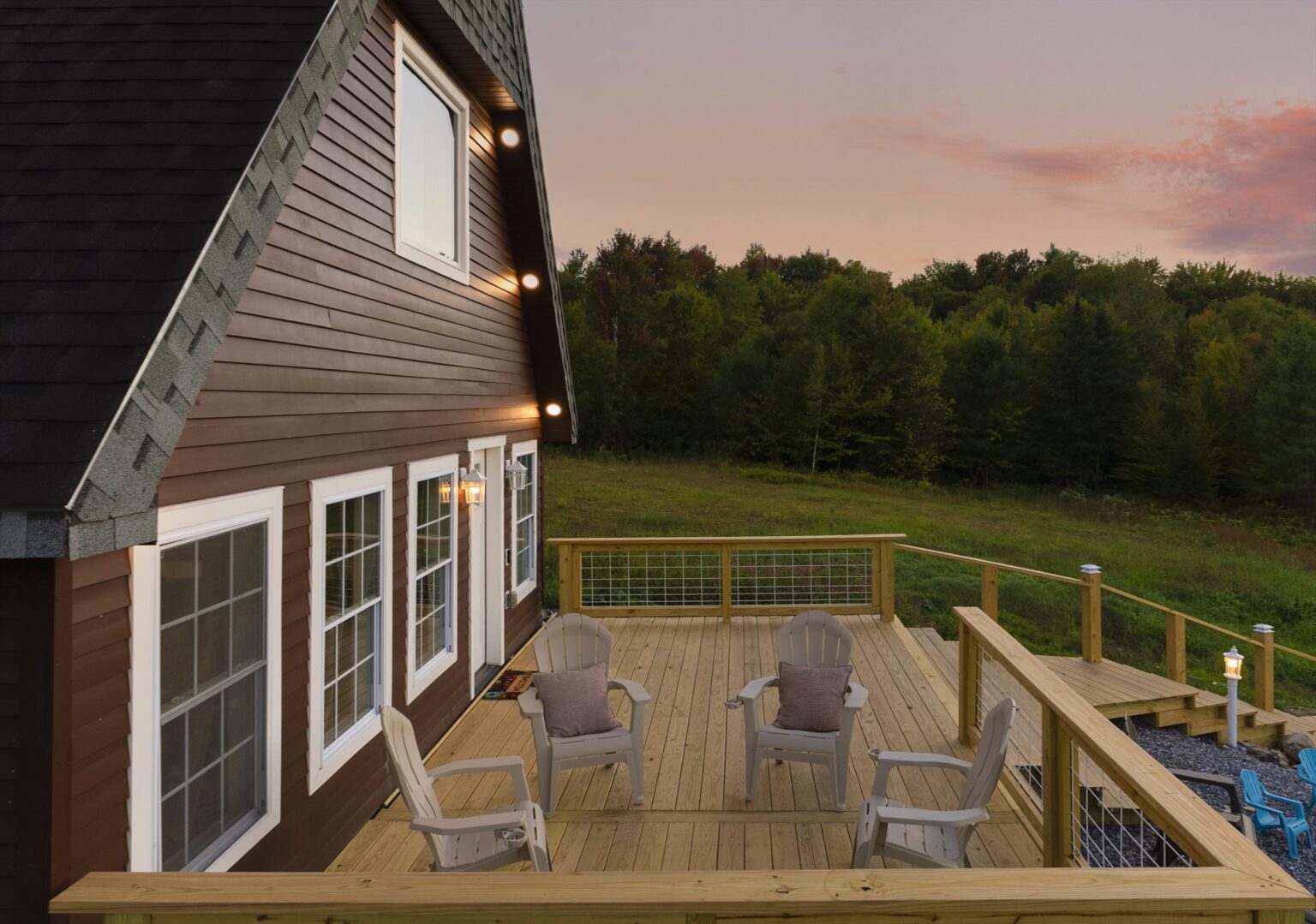 Wooden deck with four Adirondack chairs and a wire-panel railing at dusk, wall lights glowing on the siding