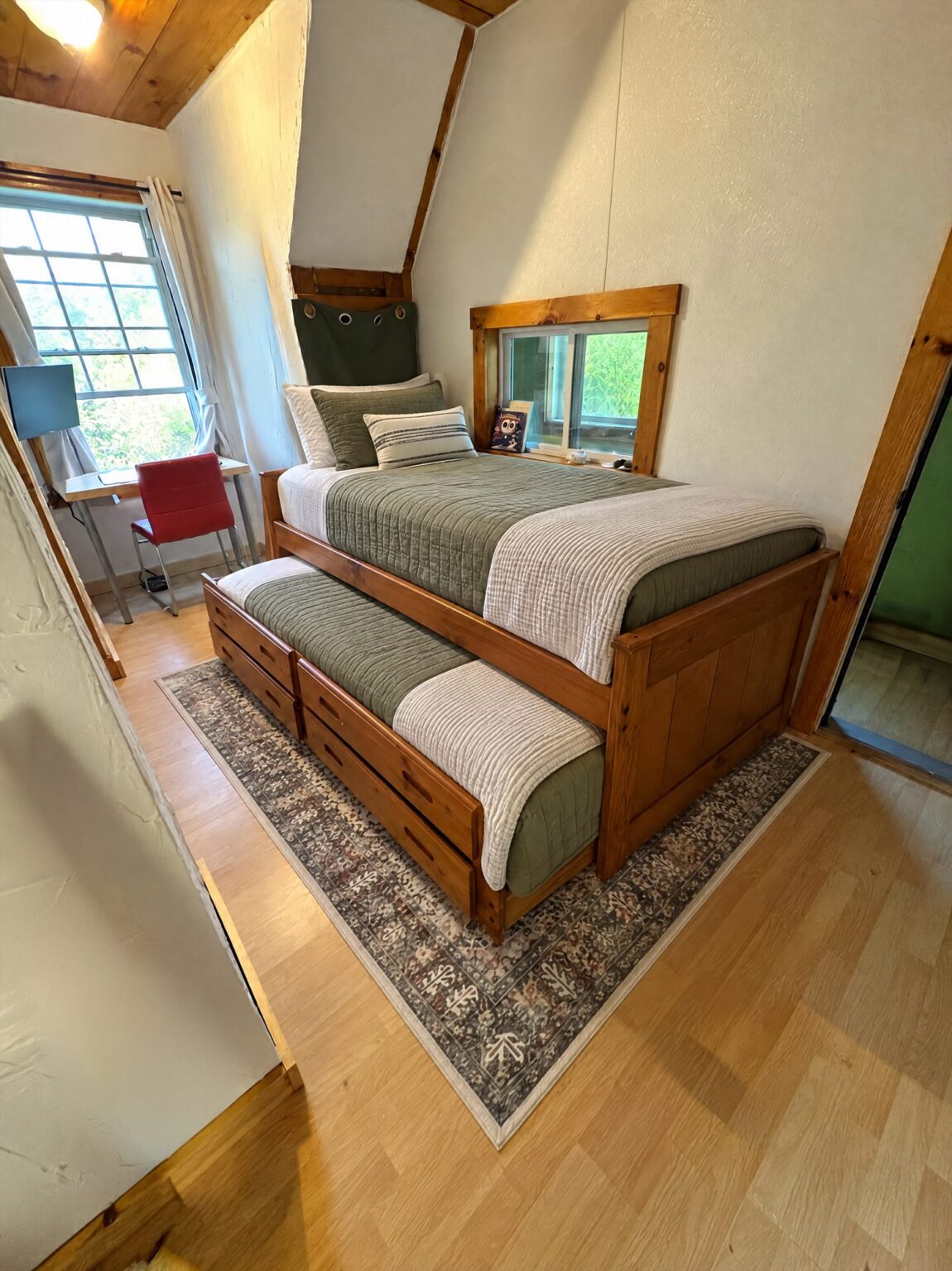 Attic bedroom with a wooden twin bed and trundle, green and cream bedding, and a desk by a window