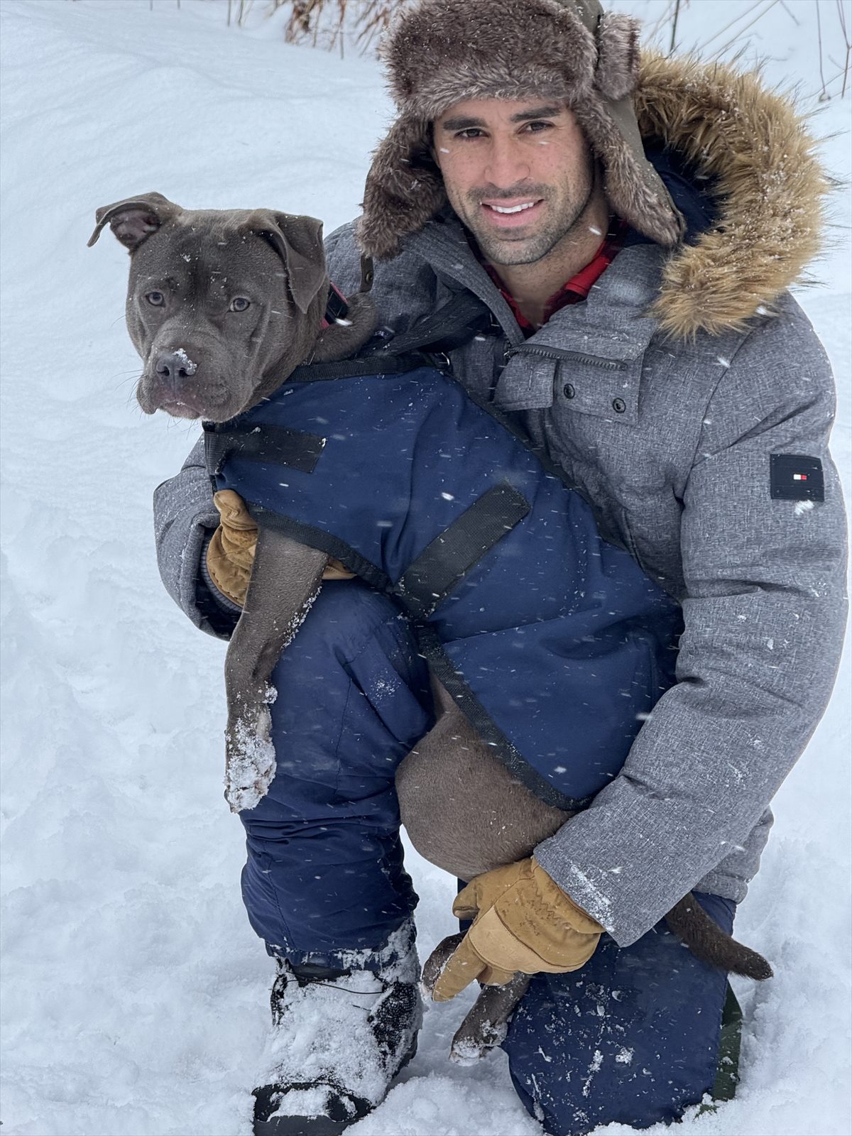 A smiling man in a fur-trimmed winter coat and hat kneeling in falling snow and holding a gray dog in a blue winter coat
