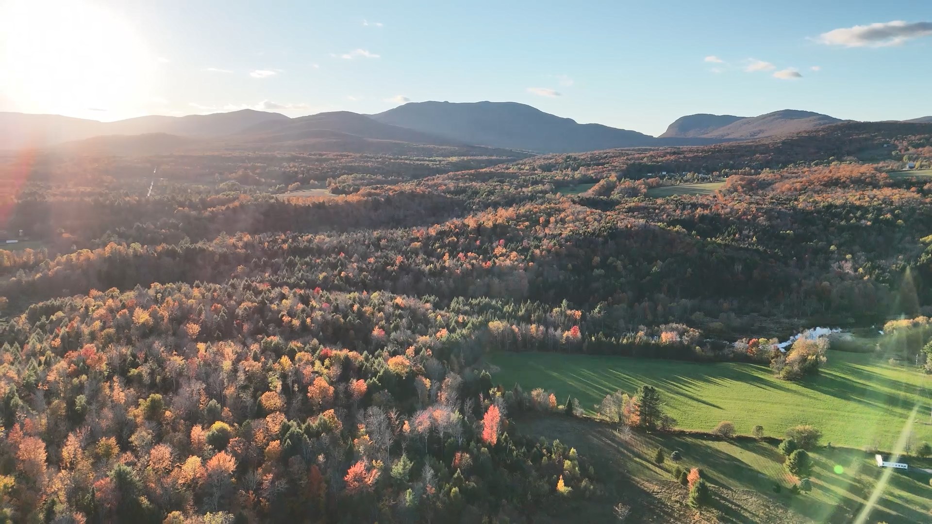 Aerial view of autumn foliage across forested hills and a green field, with mountains and a low sun in the distance