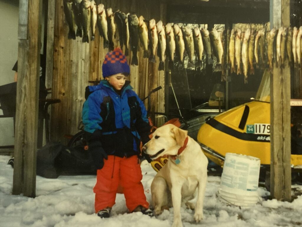 A child in a blue jacket and red snow pants stands in the snow beside a yellow Labrador, with a row of fish hanging from a shed beam behind them and a yellow snowmobile at right